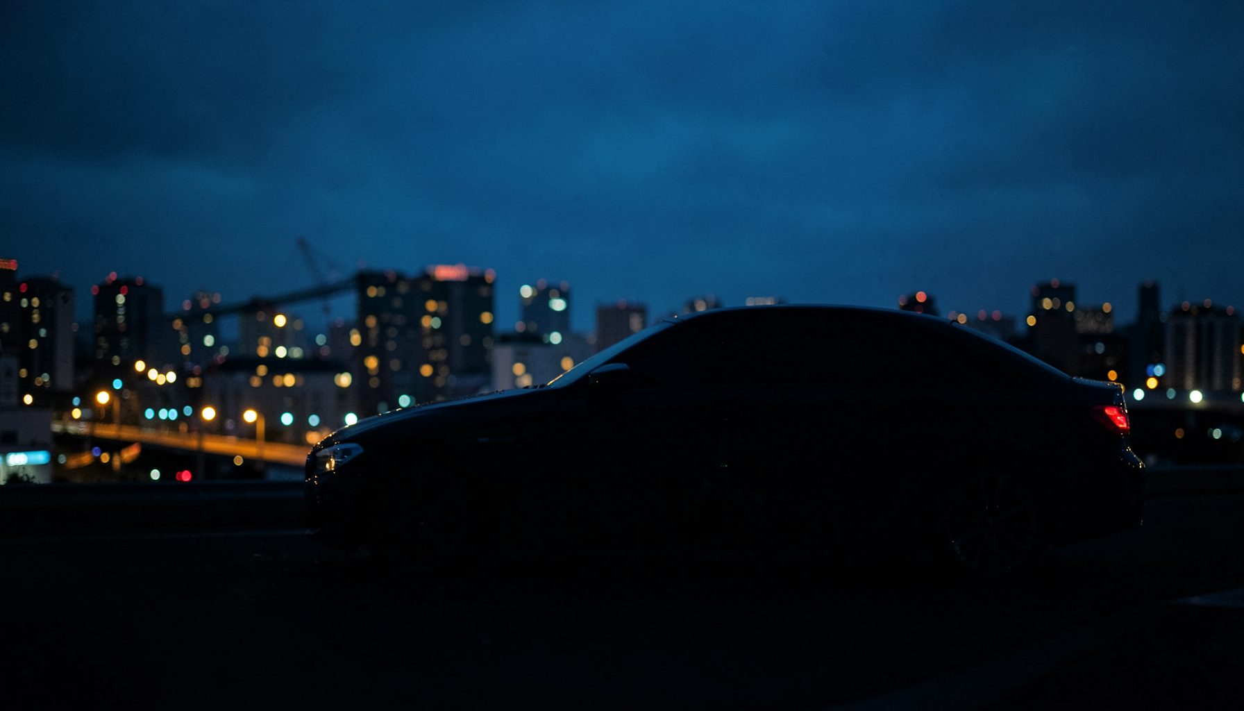 Тёмный силуэт автомобиля BMW на фоне городских огней в сумерках, глубокие синие и чёрные тона, атмосфера премиального спокойствия
