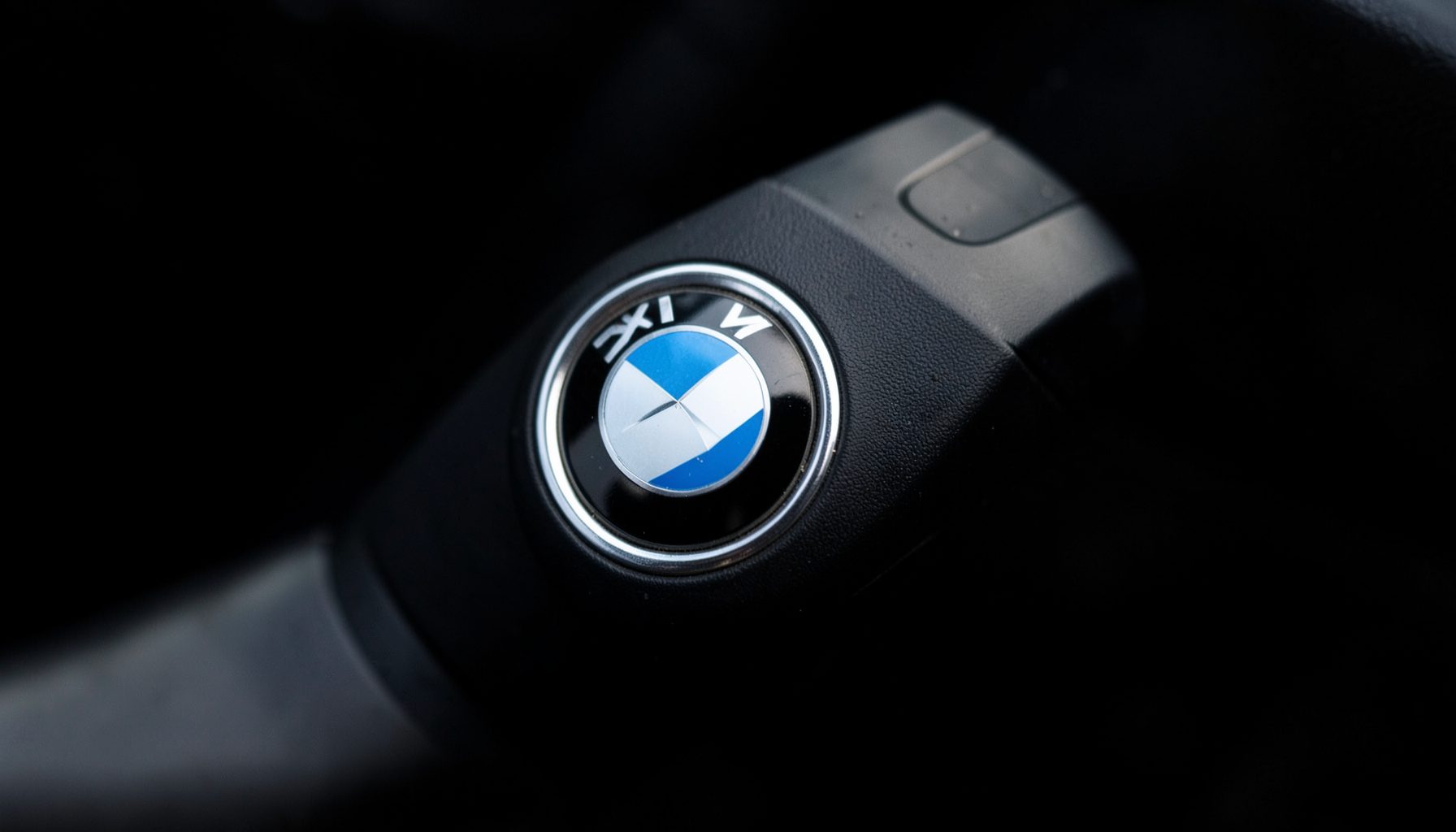 Деталь руля BMW с логотипом, крупный план, тёмный фон с мягким боковым светом, чёрные и серебристые тона